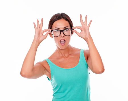 Surprised adult woman with touching her glasses with both hands while wearing her long hair tied back in a green tank top on a white backgroundの写真素材
