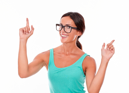 Brunette woman pointing with both hands while looking sideways at the camera and fingers up in a green tank top on a white backgroundの写真素材