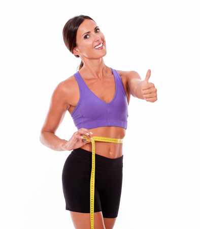 Beautiful smiling brunette gesturing thumbs up, looking at camera, while measuring her waist wearing violet and black gymnastic clothing isolatedの写真素材