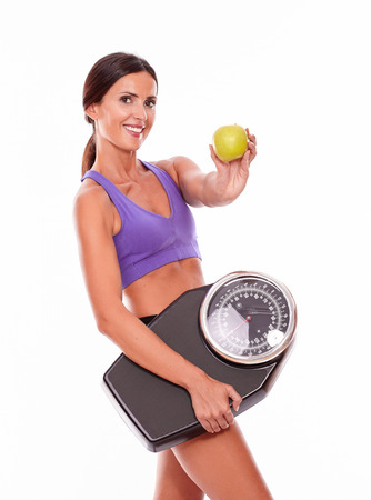 Healthy brunette woman with a scale and green apple smiling while looking at the camera with violet and black gymnastic clothing, hair tied back, isolatedの写真素材