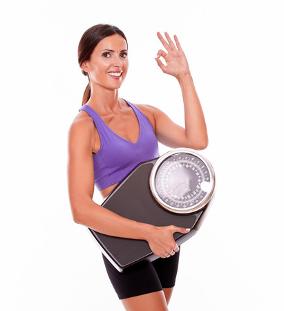 Healthy smiling brunette woman with a scale, gesturing a perfect sign while wearing her hair tied back and violet and black gymnastic clothing, isolatedの写真素材