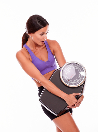 Healthy brunette woman holding and looking at a scale, while wearing her hair tied back and violet and black gymnastic clothing, isolatedの写真素材