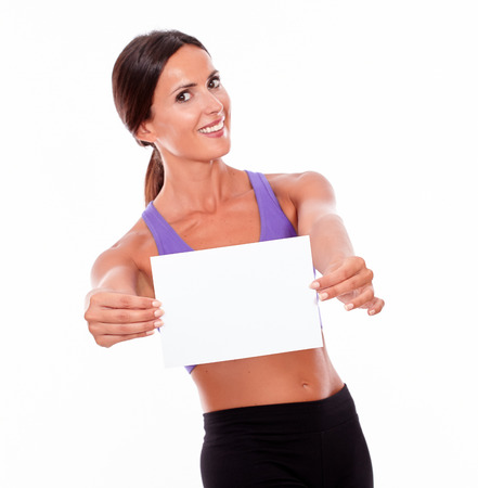 Healthy smiling brunette woman holding blank signboard in front of her with both hands looking at camera while wearing violet and black gymnastic clothing, isolatedの写真素材