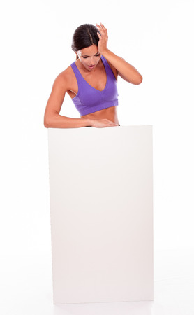 Healthy surprised brunette woman standing behind a blank placard and looking at it, hand to her head while wearing violet gymnastic clothing on white backgroundの写真素材