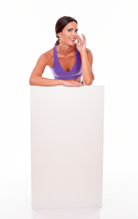 Healthy smiling brunette woman standing behind a blank placard hand to face while looking at camera wearing violet gymnastic clothing on white backgroundの写真素材