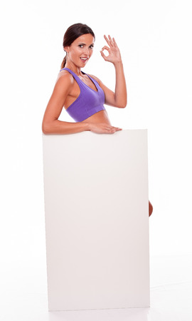 Healthy smiling brunette woman standing behind a blank placard and looking at camera, gesturing a perfect sign while wearing violet gymnastic clothing on white backgroundの写真素材