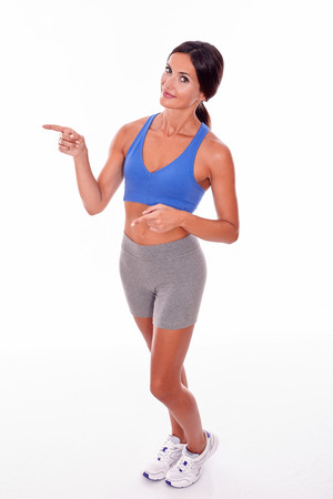 Pointing brunette woman with a toothy smile looking at the camera while wearing blue and grey casual clothing and her hair tied back, isolatedの写真素材