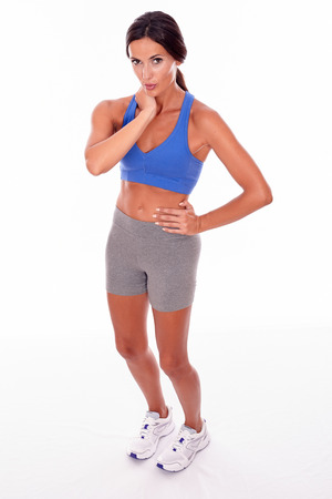 Brunette woman holding her neck with one hand and the other on her hip, looking at camera while wearing blue and grey casual clothing, isolatedの写真素材