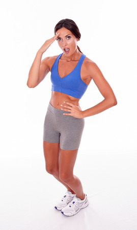 Surprised brunette woman saluting one hand to her forehead and hand on hip while looking at camera wearing blue and grey casual clothing, isolatedの写真素材