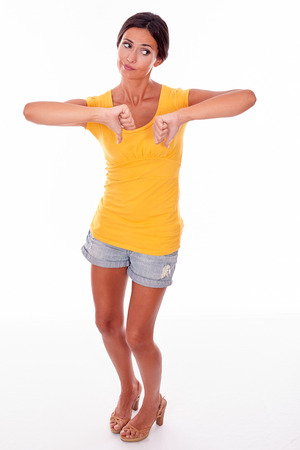 Disappointed brunette woman gesturing thumbs down in front while looking away and wearing a yellow t-shirt and short jeans isolatedの写真素材