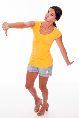 Disappointed brunette woman gesturing a thumbs down in front while looking at camera and wearing a yellow t-shirt and short jeans isolatedの写真素材