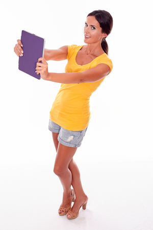 Smiling young brunette woman holding violet tablet pc while looking at camera and wearing a yellow t-shirt and short jeans on white backgroundの写真素材