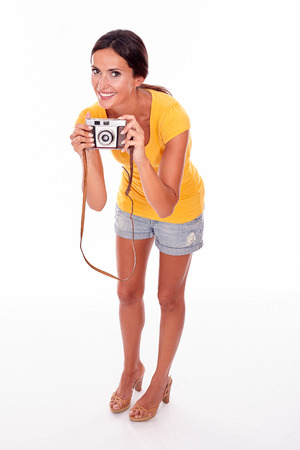 Young brunette woman with camera smiling while looking at camera and wearing a yellow t-shirt and short jeans on white backgroundの写真素材