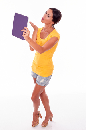 Young brunette woman holding violet tablet pc blowing a kiss at it with closed eyes wearing a yellow t-shirt and short jeans on white backgroundの写真素材
