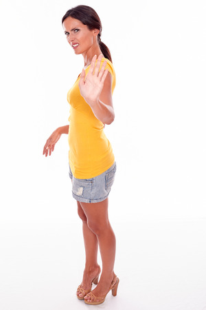 Frowning brunette woman gesturing to stop with her left hand while looking at camera and wearing a yellow t-shirt and short jeans isolatedの写真素材