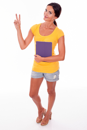 Smiling young brunette woman gesturing a peace sign while looking at camera holding a violet tablet pc wearing a yellow t-shirt and short jeans isolatedの写真素材