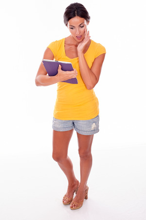 Thinking young brunette woman holding violet tablet pc while looking at tablet hand to cheek and wearing a yellow t-shirt and short jeans isolatedの写真素材