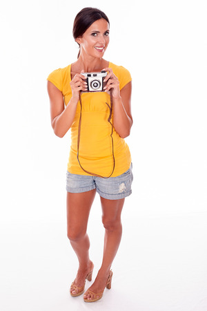 Young brunette woman with camera smiling while looking at camera and wearing a yellow t-shirt and short jeans on white backgroundの写真素材