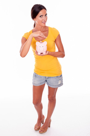 Smiling brunette holding a pink piggy bank saving coins while looking at camera and wearing a yellow t-shirt and short jeans isolatedの写真素材
