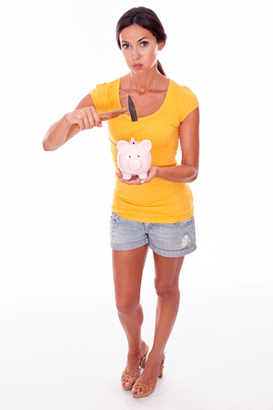 Wondering brunette holding a pink piggy bank and hammer gesturing while looking at camera wearing a yellow t-shirt and short jeans isolatedの写真素材