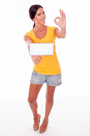 Smiling brunette woman gesturing a perfect sign with a blank envelope while looking at camera wearing a yellow t-shirt and short jeans isolatedの写真素材