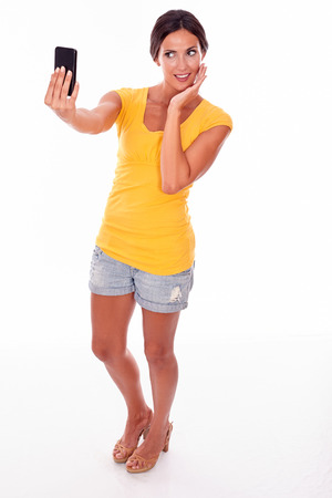 Smiling brunette holding a cell phone looking at the phone for a selfie while wearing a yellow t-shirt and short jeans isolatedの写真素材