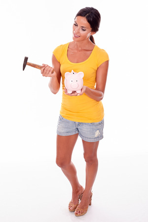 Smiling brunette holding a pink piggy bank and hammer gesturing while looking at hammer wearing a yellow t-shirt and short jeans isolatedの写真素材