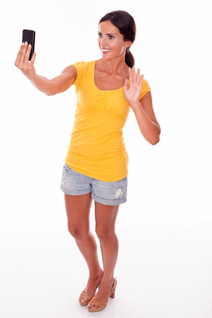 Smiling brunette holding a cell phone looking at the phone for a selfie while wearing a yellow t-shirt and short jeans isolatedの写真素材