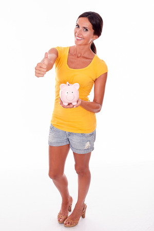 Smiling brunette holding a pink piggy bank gesturing a thumbs up while looking at camera and wearing a yellow t-shirt and short jeans isolatedの写真素材