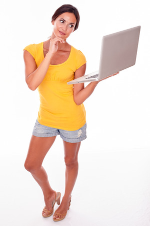 Gesturing brunette with her hand to her chin and a laptop while looking away smiling while wearing a yellow t-shirt and short jeans isolatedの写真素材