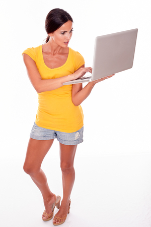 Thoughtful brunette typing on her laptop while looking at the laptop and wearing a yellow t-shirt and short jeans isolatedの写真素材