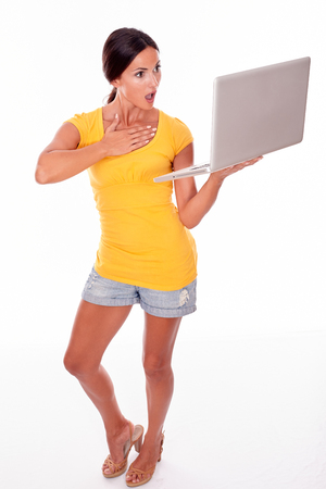 Gesturing brunette with her hand to her throat while looking at her laptop in shock while wearing a yellow t-shirt and short jeans isolatedの写真素材