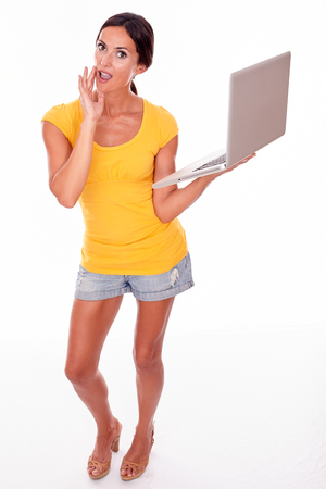 Surprised brunette with a laptop looking at the camera with her hand to her mouth while wearing a yellow t-shirt and short jeans isolatedの写真素材