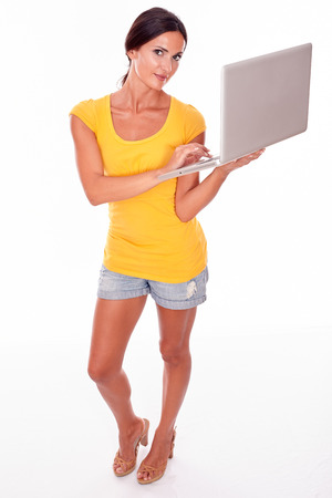Happy brunette with a laptop typing a message while looking at the camera with a smile and wearing a yellow t-shirt and short jeans isolatedの写真素材