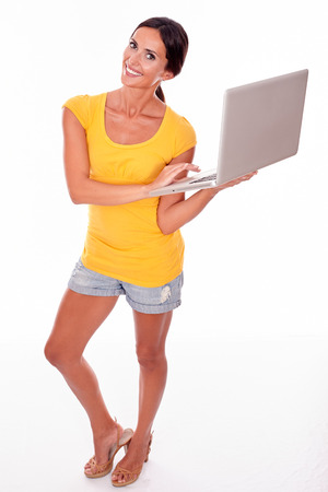 Happy brunette with a laptop typing a message while looking at the camera with a smile and wearing a yellow t-shirt and short jeans isolatedの写真素材
