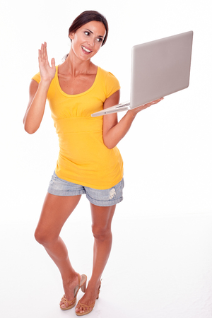 Gesturing brunette with her hand in the air while looking at her laptop with a smile, wearing a yellow t-shirt and short jeans isolatedの写真素材