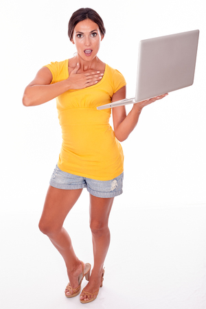 Gesturing brunette with her hand to her throat and a laptop while looking at camera in shock, wearing a yellow t-shirt and short jeans isolatedの写真素材