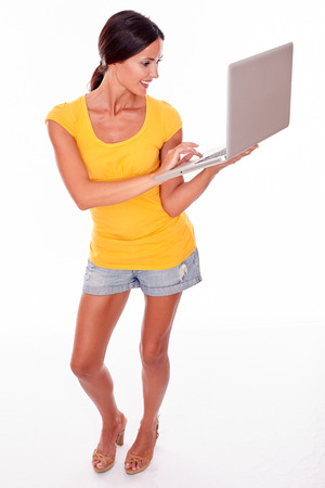 Smiling brunette with a laptop typing a message while looking at laptop with a smile and wearing a yellow t-shirt and short jeans isolatedの写真素材