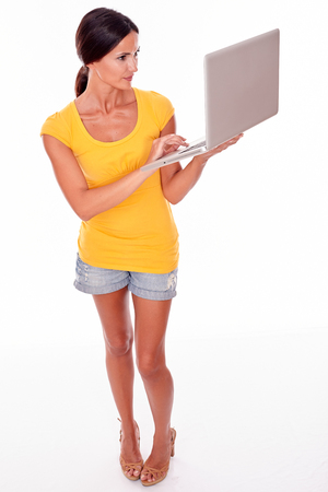 Smiling brunette with a laptop typing a message while looking at laptop with a smile and wearing a yellow t-shirt and short jeans isolatedの写真素材
