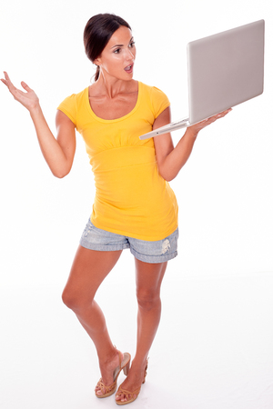 Gesturing brunette with her hand in the air while looking at her laptop and wearing a yellow t-shirt and short jeans isolatedの写真素材