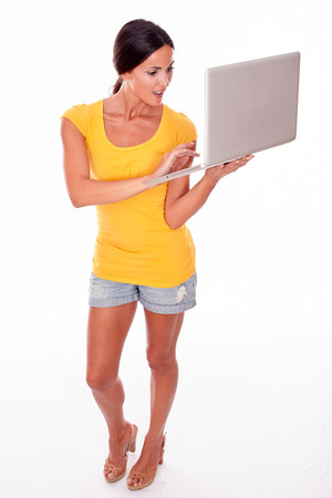 Surprised brunette with a laptop typing a message while looking at the laptop open mouthed and wearing a yellow t-shirt and short jeans isolatedの写真素材