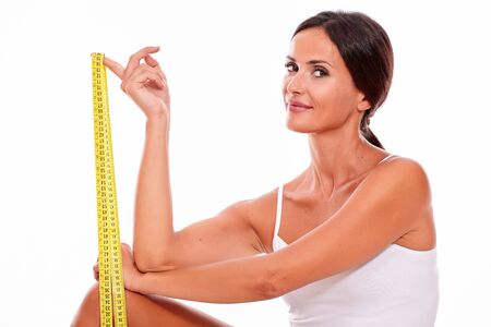 Young smiling brunette woman showing a tape measure looking at camera thoughtfully while wearing a white tank top and her hair tied back, isolatedの写真素材