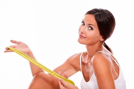 Young brunette woman showing a tape measure looking at camera with a contemplating smile wearing a white tank top and her hair tied back, isolatedの写真素材