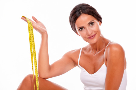 Young brunette woman showing a tape measure looking at camera with a smile wearing a white tank top and her hair tied back, isolatedの写真素材