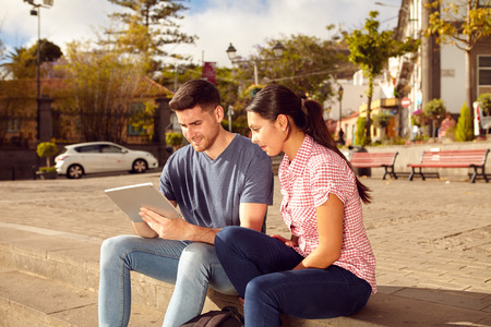 Young couple sitting in a town square on a bench in the sunshine looking at a tablet while dressed casually in jeans and t-shirtsの写真素材