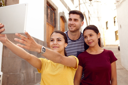 Young tourist couple on vacation with a backpack taking a selfie with a friend in a narrow street while dressed in casual clothesの写真素材