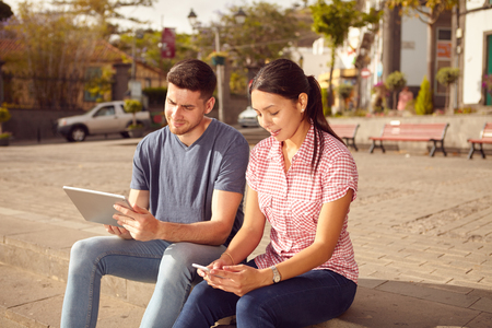 Young couple sitting in a town square on a bench looking at a tablet and a cell phone while dressed casually in jeans and t-shirtsの写真素材