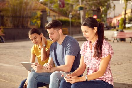 Three young people sitting in a town square on a step looking at a tablet and a cell phone while dressed casuallyの写真素材