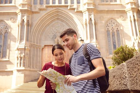 Young tourist couple on vacation with a backpack, looking at a map while dressed casually in jeans and t-shirts with a cathedral behind themの写真素材