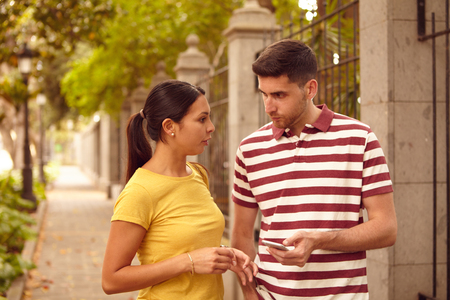 Young couple looking at each other and talking with a cell phone and a hand in his pocket while dressed casually in jeans and t-shirtsの写真素材